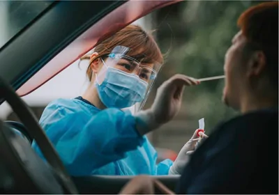 Woman in face shield and blue gown taking cotton swab of patient's mouth while patient sits inside of car Woman in face shield and blue gown taking cotton swab of patient's mouth while patient sits inside of car